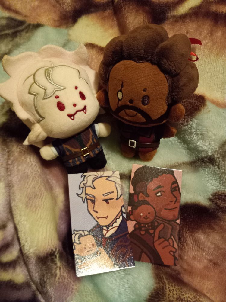 Chibi Astarion and Wyll plush with photocards made by @miyabau. They are wearing their base armor from when you first meet them in the game.

The two photocards have their larger selves smiling down at their plush forms.