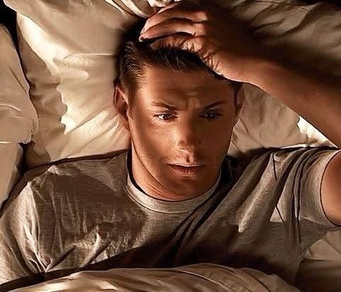 Jensen Ackles laying in bed with one hand on his head. His expression looks agitated.