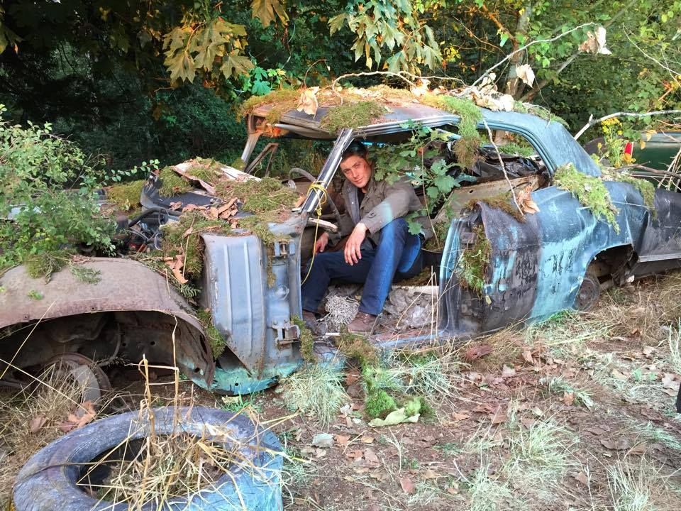 Jensen Ackles sitting in an old, damaged car that is covered with moss and dirt