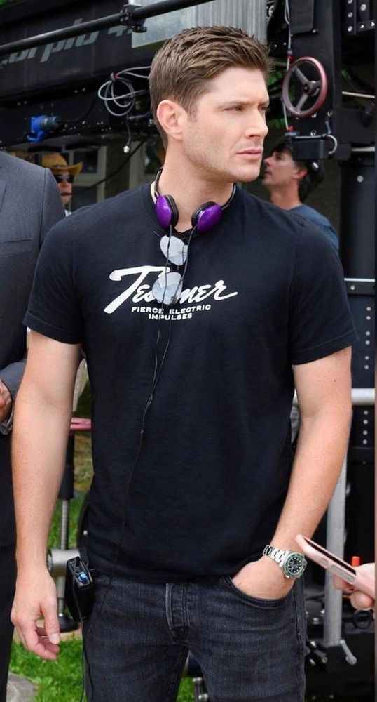Jensen Ackles dressed in black and standing with headphones around his neck. One hand is in his pocket