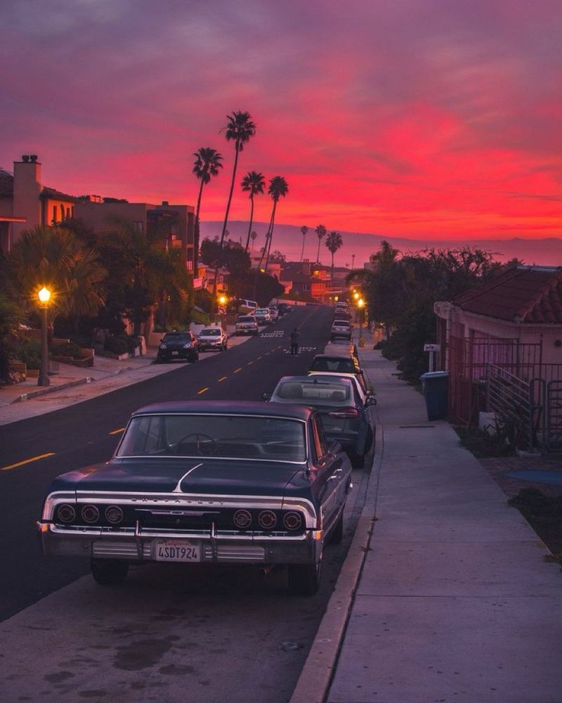 A reddish-orange sunset looms above a street at dusk. Cars are parked along the side of the street & palm trees tower in the distance.