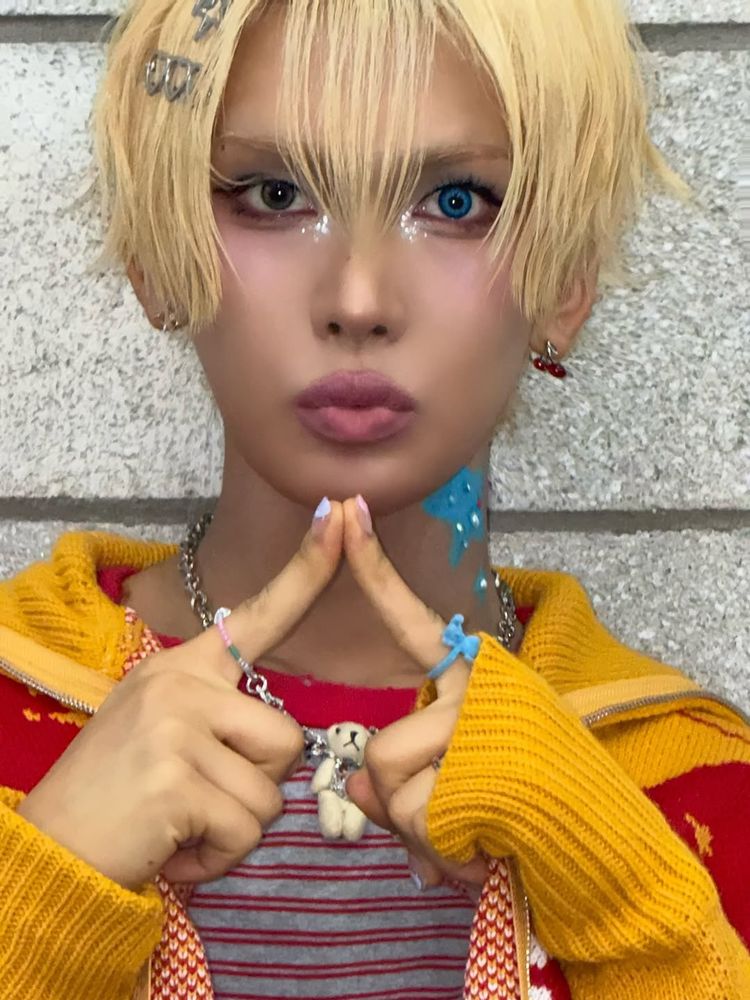 Hyun with blonde hair pouting at the camera. He’s holding the tips of his pointer fingers together under his chin.