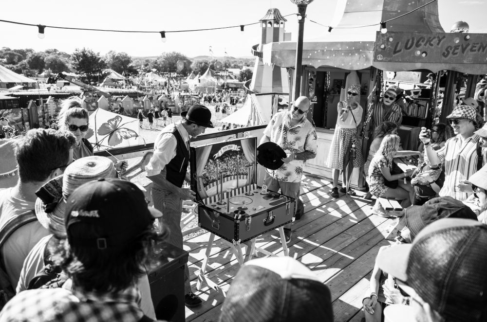 a man in a bowler hat shows off a flea circus to a crowd.