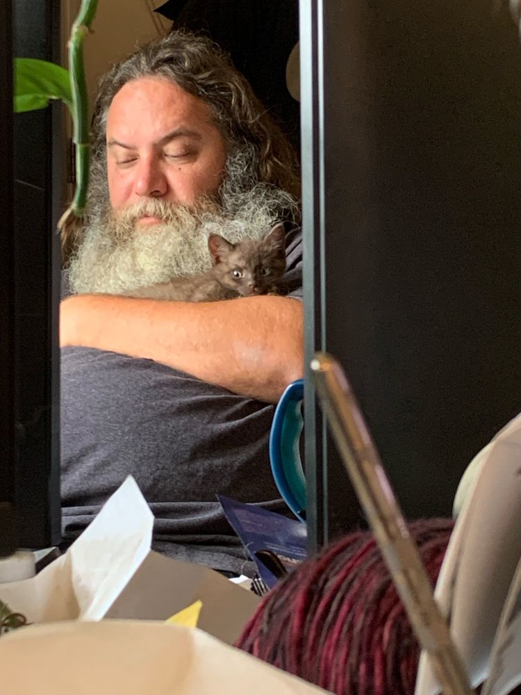 A small kitten peers at the camera from the arms of a bearded man