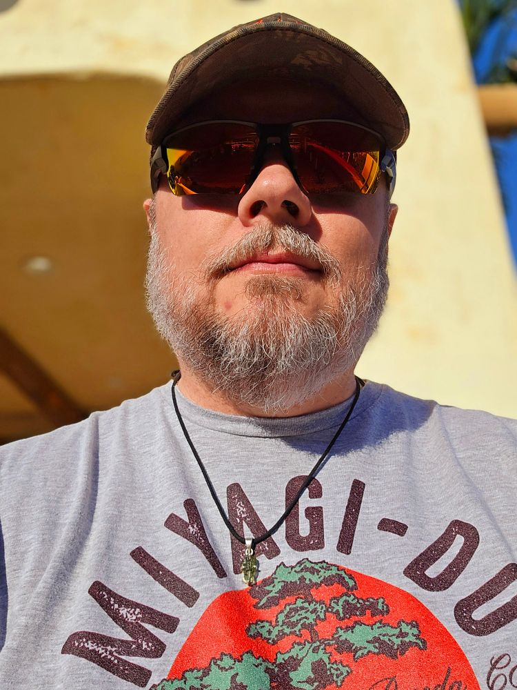 Cis man with a beard wearing a grey t-shirt, orange sunglasses, and a green camo hat.