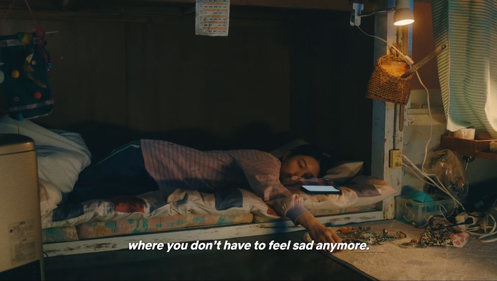 Continues from previous: "where you don't have to feel sad anymore." It's now a wide shot of the girl ont he bed. She's reaching for a keychain from a pile that's lying on a low table next to her bed.