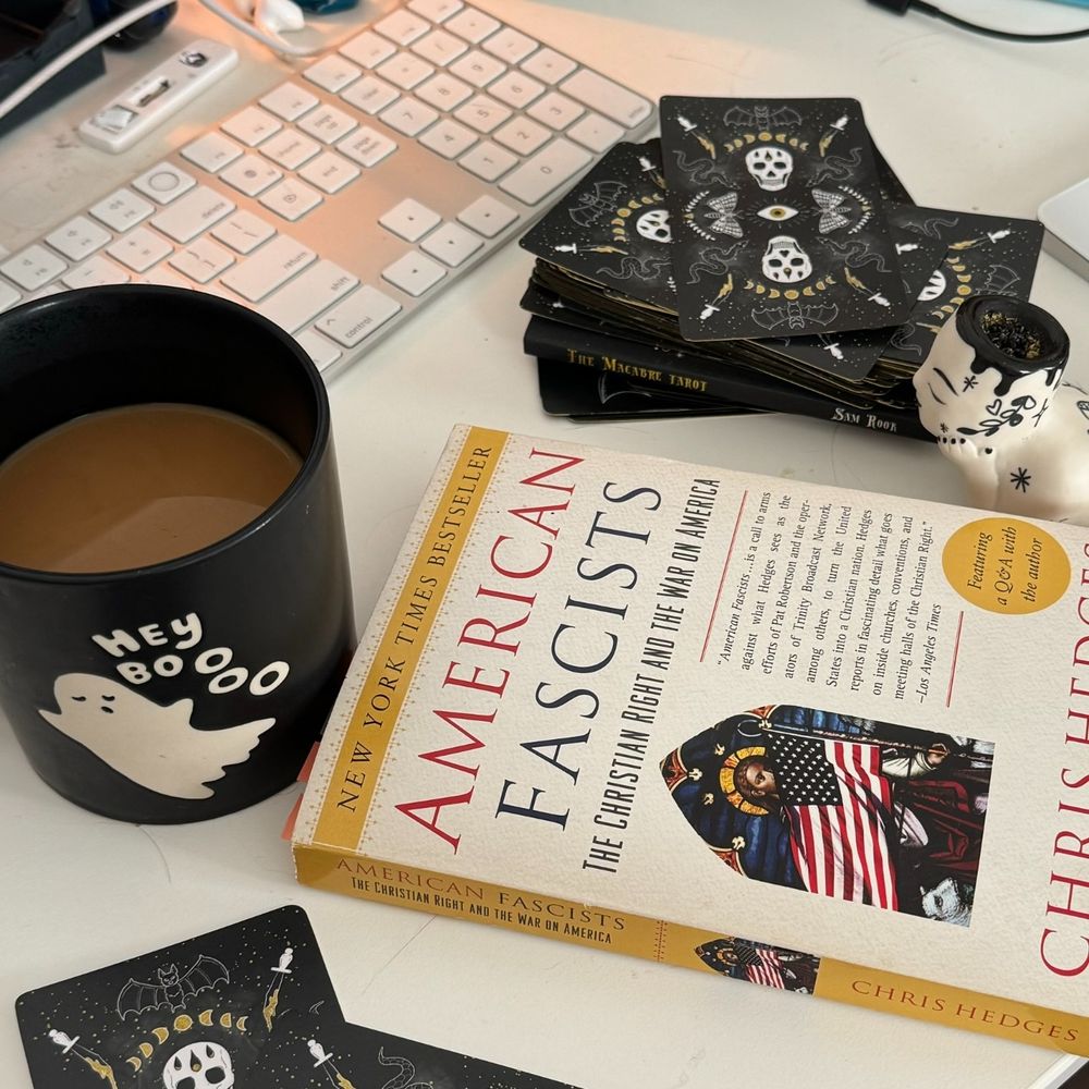 Snapshot of a writer's desktop with Halloween-themed cards from "The Macabre Tarot," magically-brewed coffee in a "hey boooo" ghost mug, a pipe for wake and bake, and the book "American Fascists: The christian right and the war on america" by Chris Hedges.