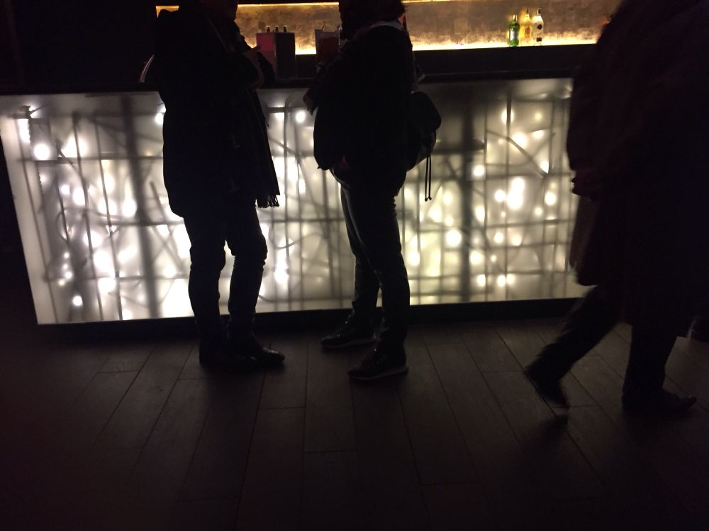 Von hinten mit Lichterketten beleuchteter Bartresen, davor Menschen, von denen nurSilhouetten erkennbar sind 