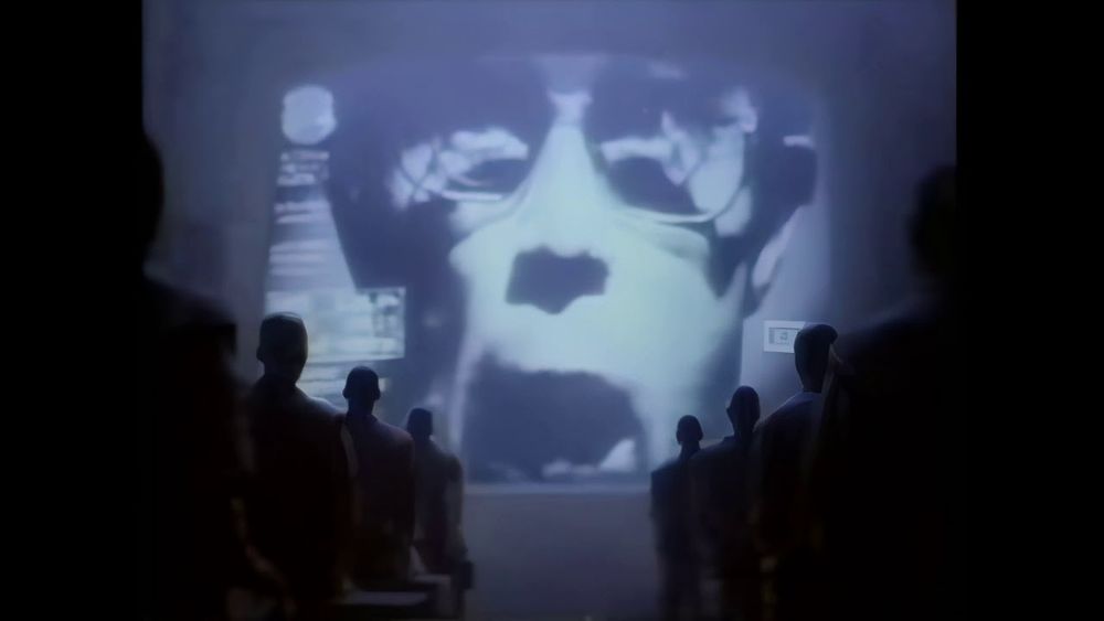 A screen capture of Apple’s famous 1984 Super Bowl commercial. It shows an audience forced to watch the authoritarian leader’s hate message. 