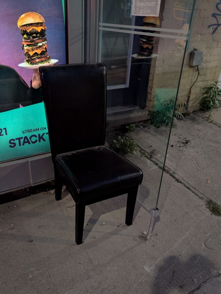 A black leather armless chair, slightly scuffed, sits in the corner of a transit stop shelter, against a glass wall. Behind it, on a glowing advertisement poster, a hand holds up a tall overly stacked cheeseburger.