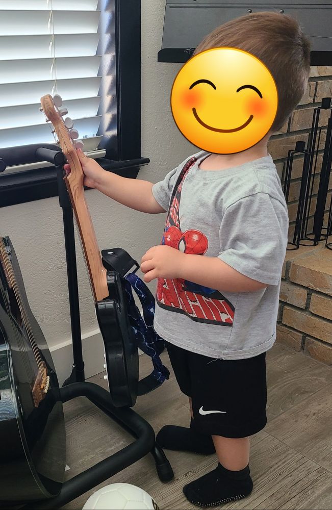 A toddler plays with his child-sized toy guitar during free play time. His identity is shielded with a smiling face sticker.