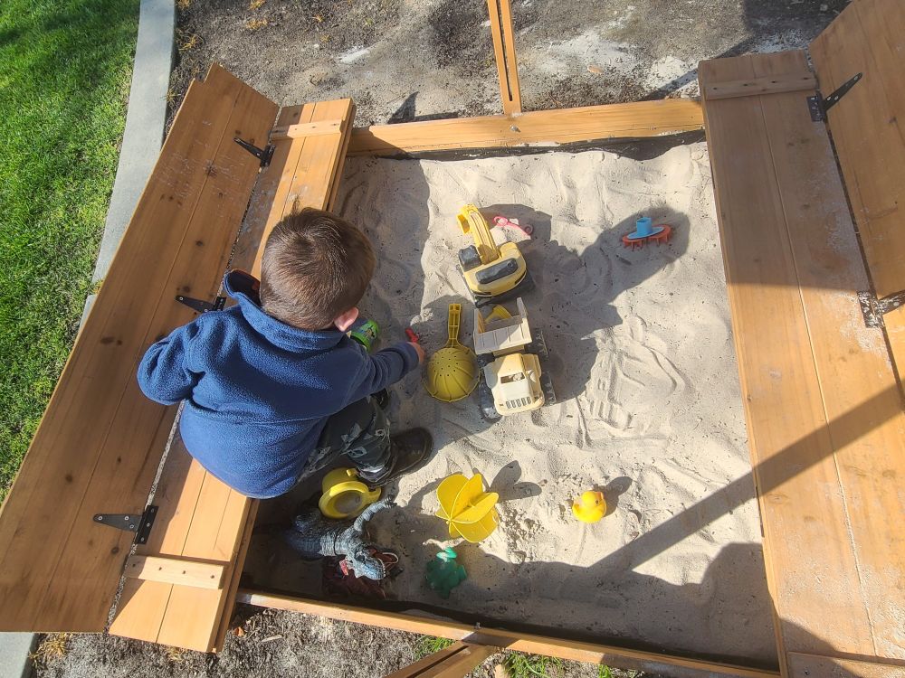The toddler boy that I babysit is shown playing in a sandbox with various sand toys.