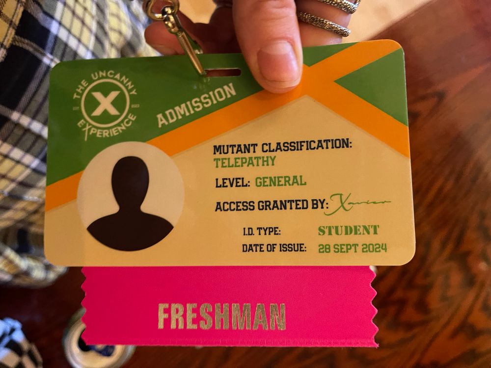 A picture of event badge reads: mutant classification-telepathy, level-general, access granted by-Xavier, I.D. type-student, date of issue-28 Sept 2024. A pink badge ribbon attached to badge reads “freshman”