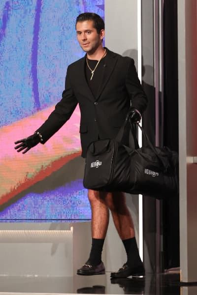 A man wearing a black t-shirt, black coat, black gloves, black socks, black shoes, and no visible shorts or pants, as he's also carrying a black duffle bag