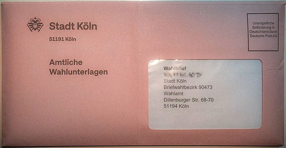 Das Foto im Querformat zeigt ein rosa Briefkuvert im Format DIN lang. Es hadelt sich um amtliche Wahlunterlagen der Stadt Köln. Adressiert ist der Brief an den zuständigen Briefwahlbezirk.