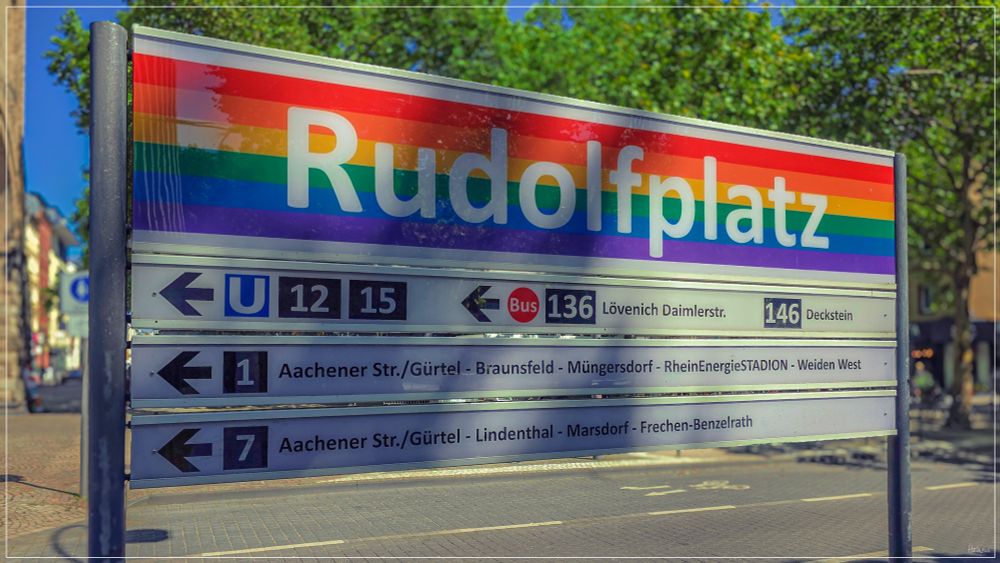 Das Colorfoto ist im Querformat angelegt und zeigt in Großaufnahme das Schild der KVB-Haltestelle Rudolfplatz in Köln. Der Schrift ist mit den Regenbogenfarben hinterlegt und steht symbolisch für die Weltoffenheit der Stadt Köln.