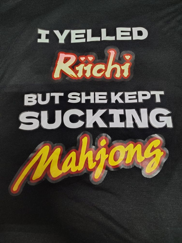 A black shirt that reads "I yelled Riichi but she kept sucking Mahjong"