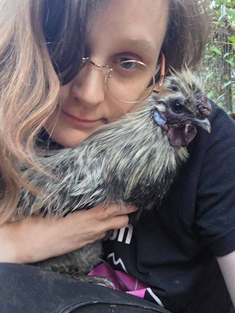 Focus on a crouching human holding a chicken to their chest. Both are looking at the camera. The human is pale-skinned and feminine, with long asymmetrical dirty blonde hair and gold circle-rimmed glasses. The chicken is a small speckled black-and-silver silkie rooster The roo's feet have purchase on the human's legs, and the human is holding the roo with good support of its upper body. They are tight enough to feel each other's warmth. The human is tilting their head and smiling, with their cheek to the roo.