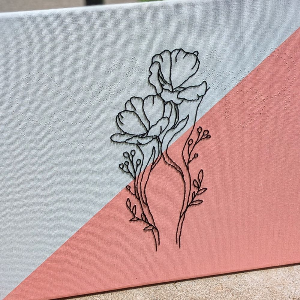 Close up of a line art embroidery on canvas WIP. The background is painted half white and half pink on a diagonal. The stitches are black. The design is flowers in the shape of a uterus. Only the middle portion is complete, but you can see the holes poked in the canvas for what will represent the fallopian tubes and ovaries.