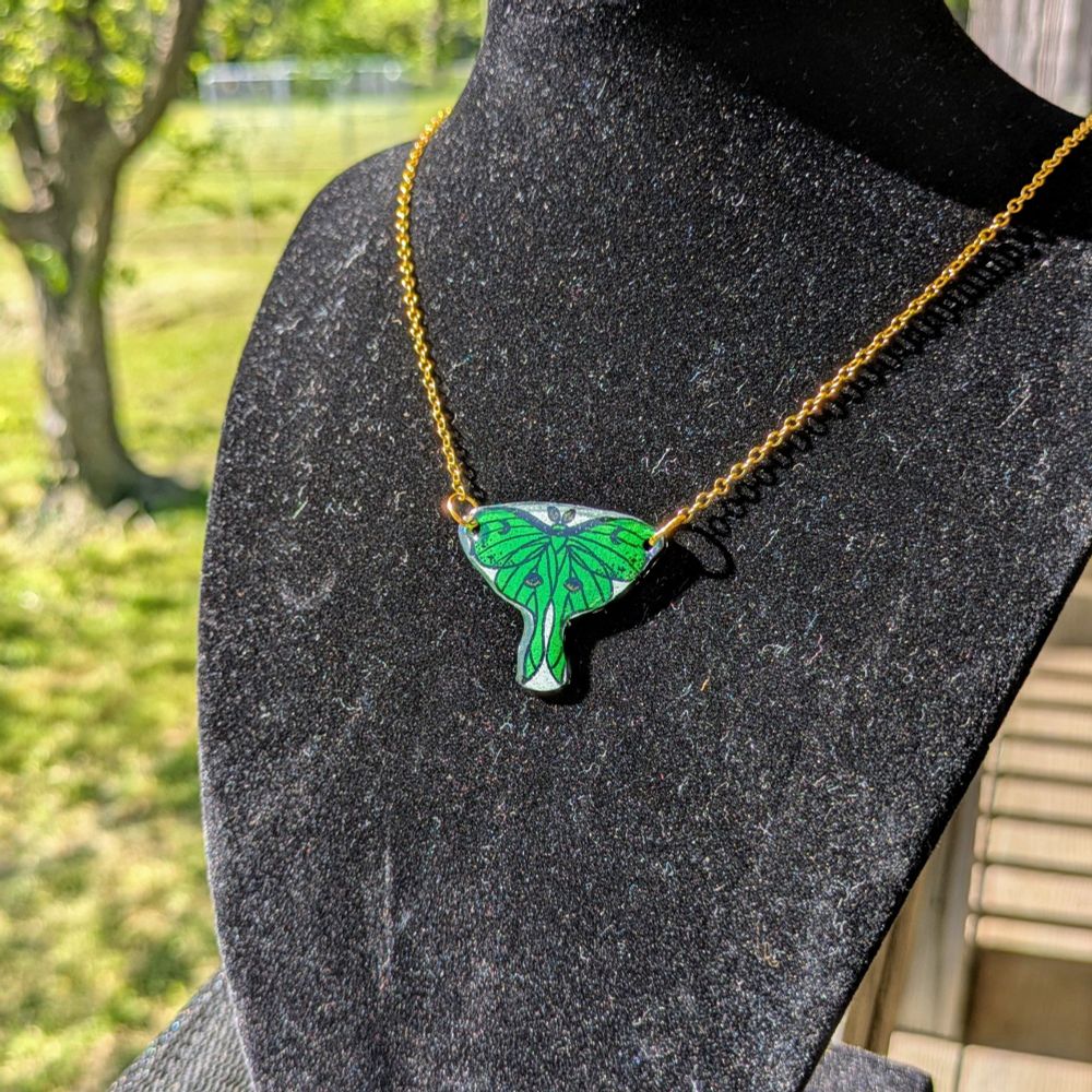 A necklace on a black display bust with green nature in the background. The necklace is a Luna Moth with a golden chain attached to each wing.