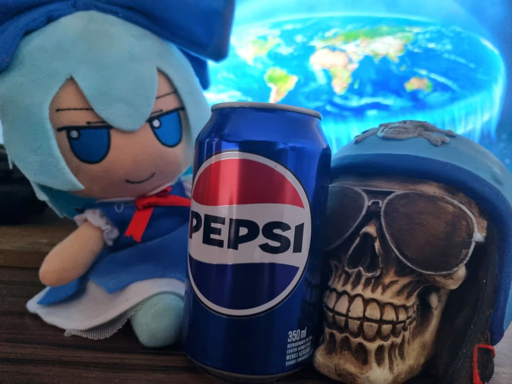 Uma pelúcia Fumo da Cirno ao lado de uma lata de refrigerante Pepsi e uma escultura de caveira. Ao fundo tem uma representação de uma Terra plana. 