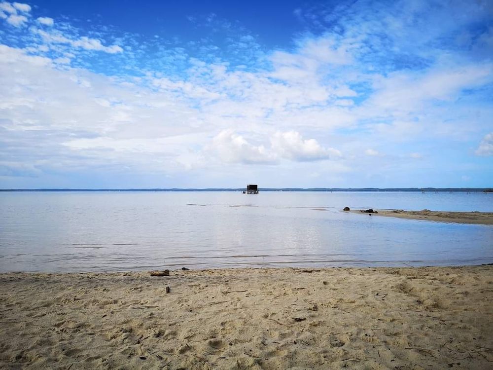 Plage de sable, ciel bleu avec nuages blancs, du sable en premier plan, l'eau du lac derrière et au fond on aperçoit une cabane de pêche 