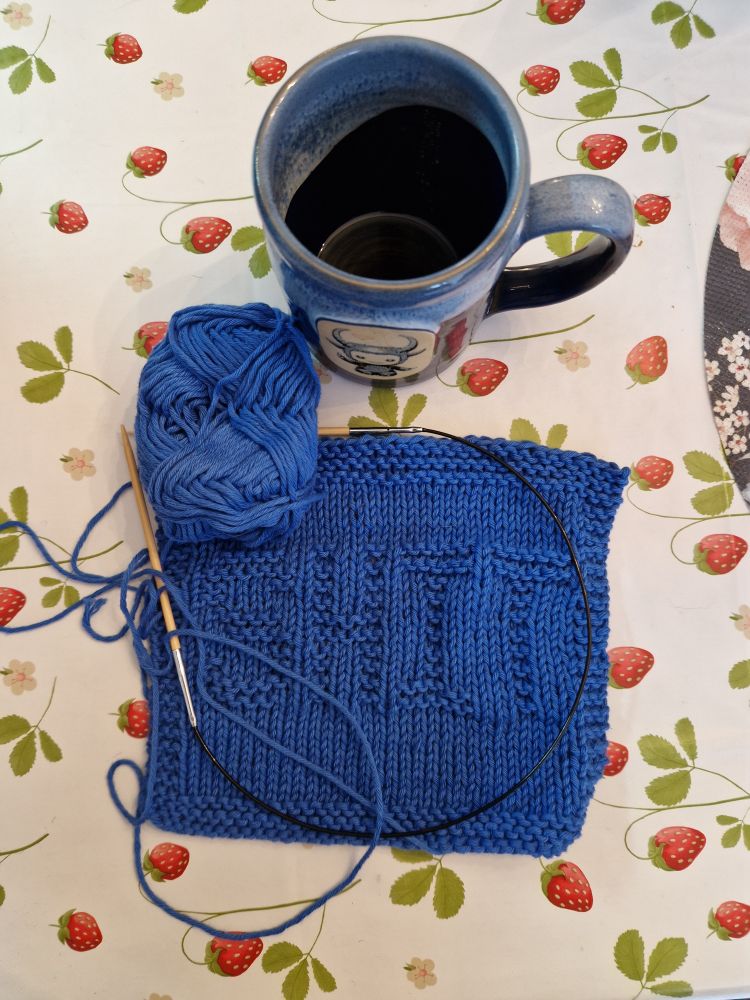 Blue knitted dishcloth with the word "shit" purled. Blue mug of coffee with baby baphomet on it. White tablecloth with 🍓 and leaves.