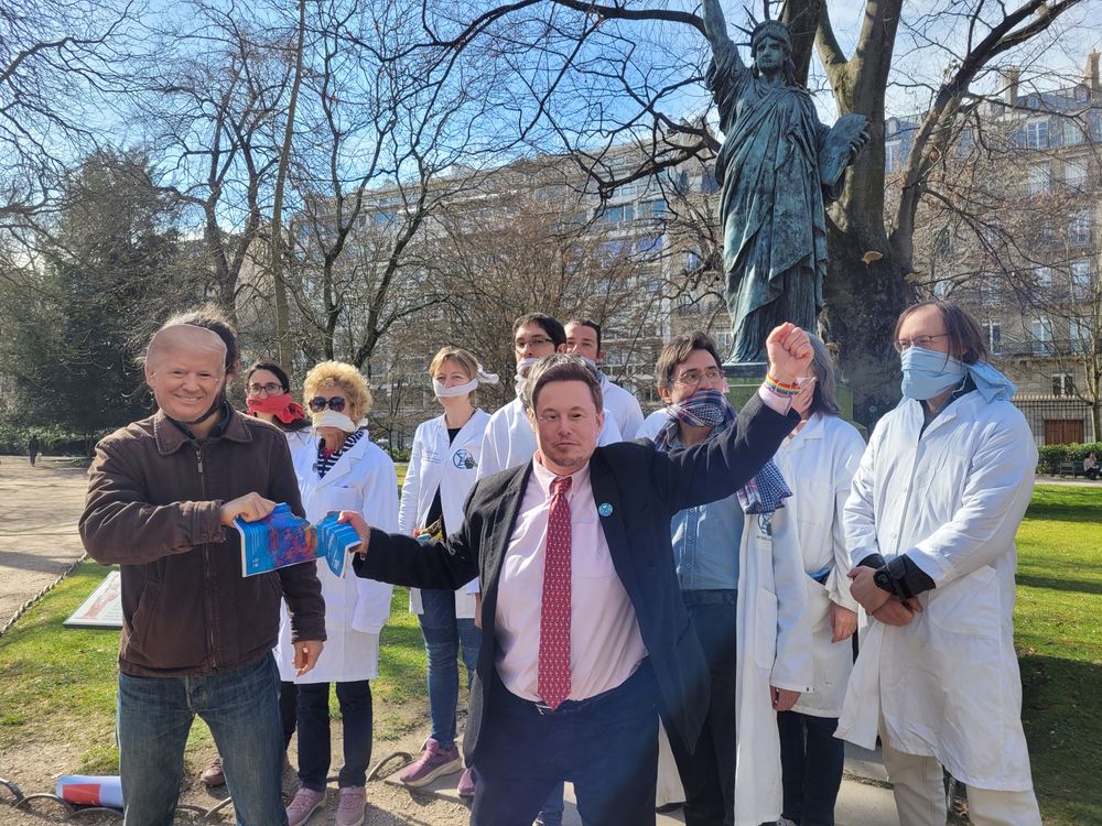 Deux personnes avec des masques de Trump et Musk déchirent un rapport du GIEC devant un groupe de scientifiques bâillonnés, aux pieds de la statue de la liberté, dans le jardin du Luxembourg.