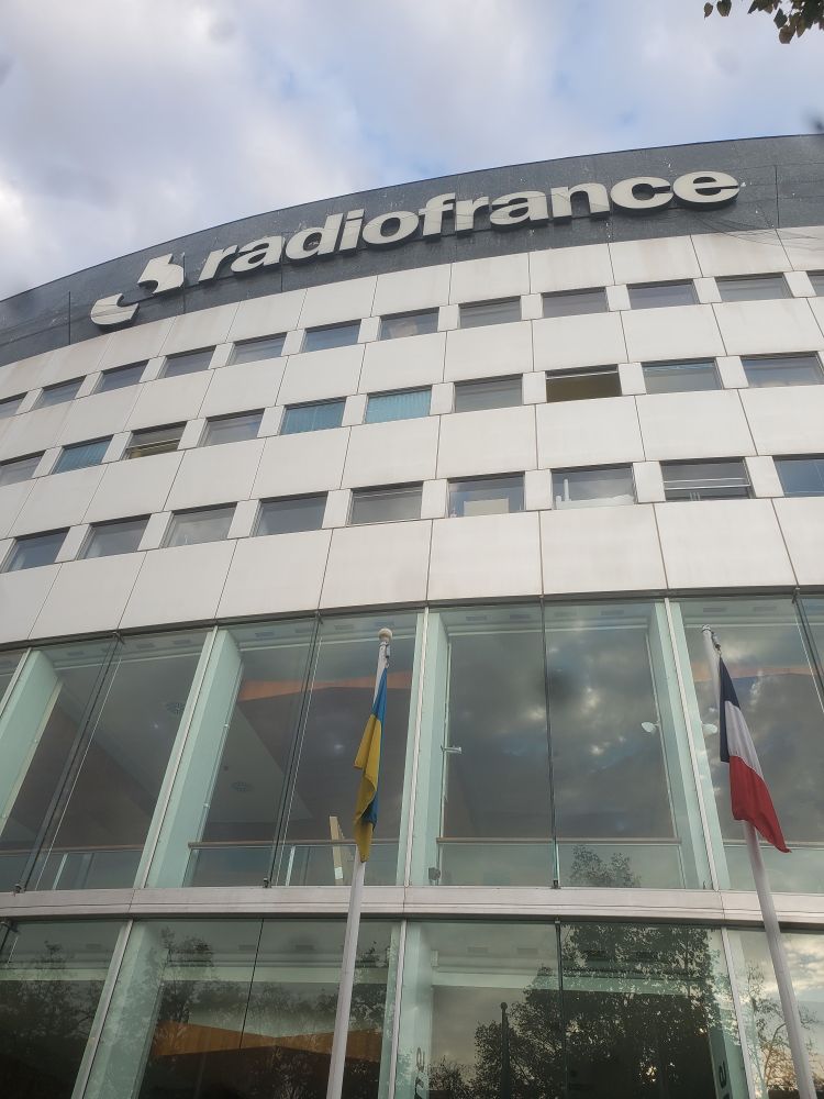 Photo d'un grand bâtiment moderne avec des hautes vitres et des drapeaux et marqué Radiofrance en haut 