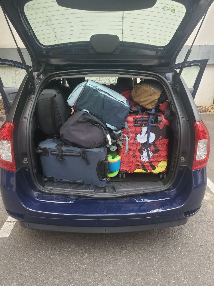 Photo d'un grand coffre de voiture plein de bagages, on voit 2 valises sur le devant dont 1 valise Mickey et pas mal de bazar sur le dessus avec des sacs à dos qui dépassent 