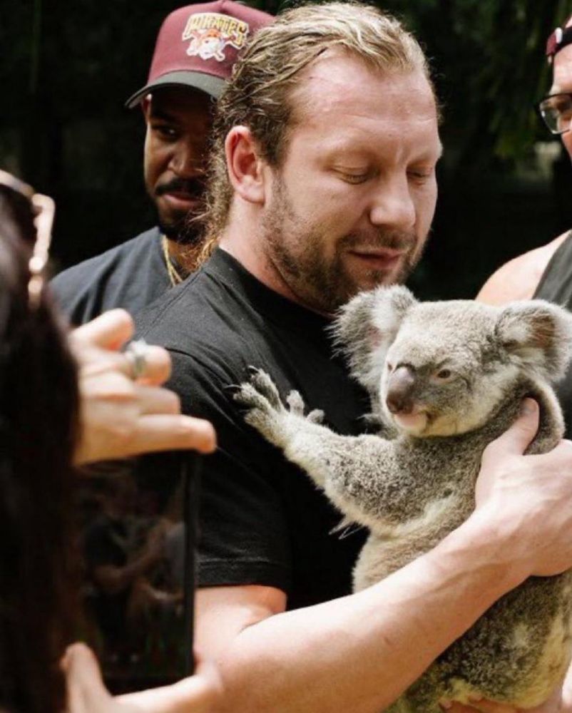 Kenny Omega in a black shirt holding a koala. It’s grey and looks slightly toward the camera as it holds onto Kenny’s chest. Kenny has long hair that is pulled back and a slight beard. 