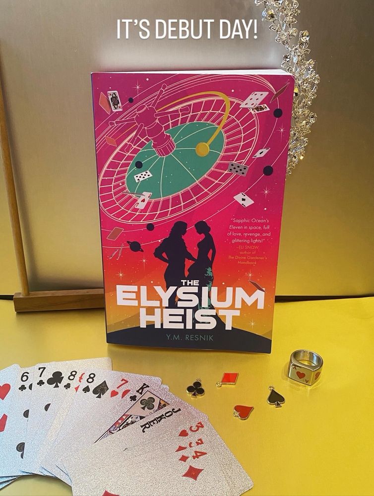 Shiny gold floor and shiny silver wall. On the bottom left is a fanned deck of silver playing cards. On the bottom right is a set of charms in the shape of ace, spade, heart, and Diamond and a silver ring with an etched ace of hearts. Propped up against the wall is a copy of The Elysium Heist. Leaning against the book is a dice stick. Behind them stretched across the wall is a Diamond tiara. 