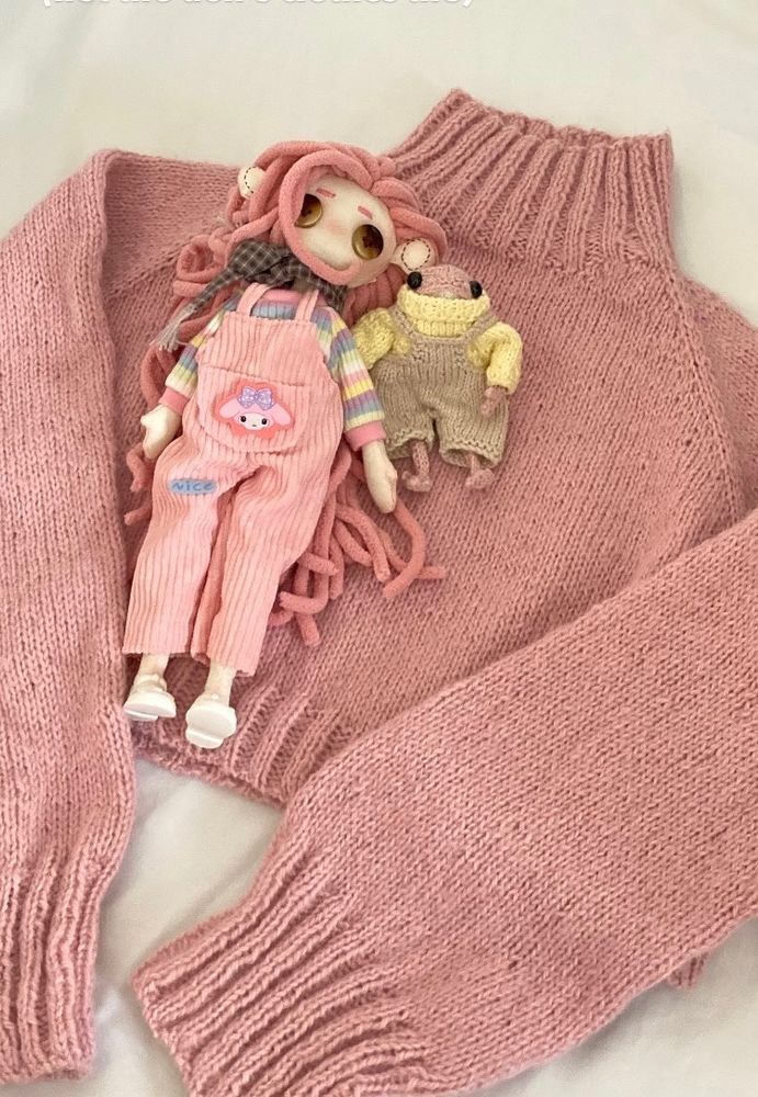 3 handmade items: a fabric doll with pink hair and clothes (the later not made by me), a knitted pink frog with a light yellow jumper and beige overalls. they are both on a pink knitted jumper with ribbed neck and cuffs. everything is very cute and pink.