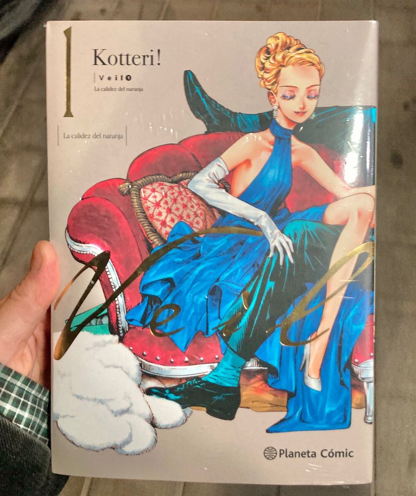 a hand holding the manga. the title is gold and behind it’s the blonde protagonist with her eyes closed, dressed in a blue dress and seating on a red sofa. there are two legs peaking from the right, indicating it’s a part of a bigger illustration. 
