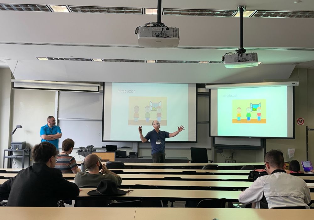 Two professors stand in front of a university lecture hall. They are presenting a student's paper. The projector behind them displays a slide titled "Introduction" with a photo of three cartoon boys on it.