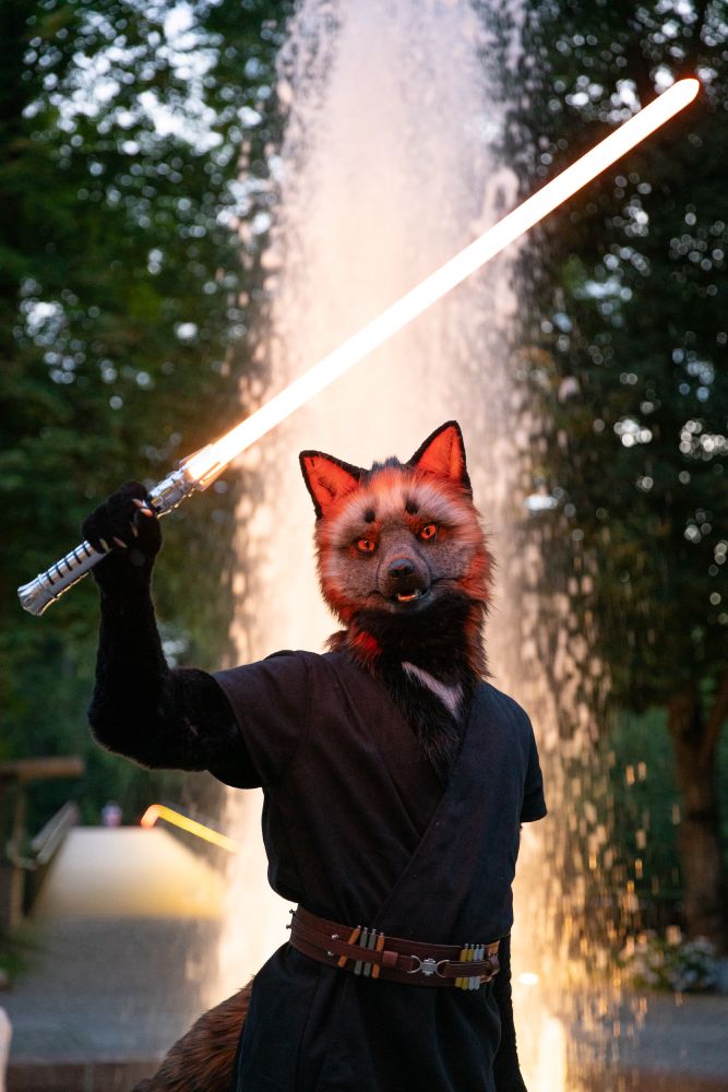 Veylan, an anthropomorphic cross fox (fursuit), standing in front of an illuminated fountain at dusk. He is wielding a lightsaber, holding it up as if deflecting an incoming attack.
