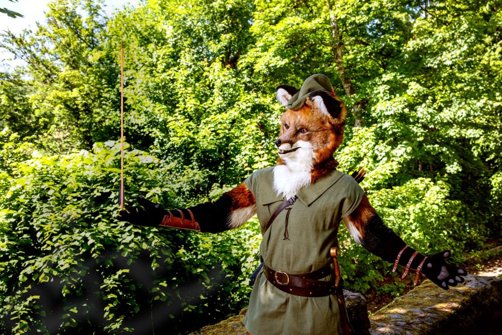 Re-enactment of an iconic scene from Disney's Robin Hood: Arikh (realistic red fox fursuit) cosplaying Disney's Robin Hood, standing in front of a moss-covered wall in the forest, casually holding out his arm and balancing an arrow on the pad of his index finger.