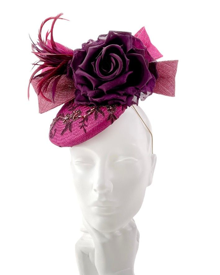 A rich berry-toned perching pillbox hat with embroidered beading, plum silk rose, sculpted bow and feather detail. Displayed on a white mannequin head. Designed as an elegant wedding hat for the Mother of the Bride and Mother of the Groom, perfect for a Christmas or winter wedding.
