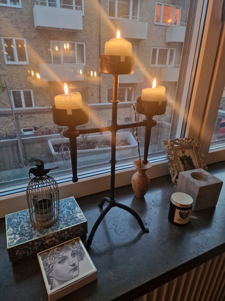 A photo of a stone windowsill with a medieval style iron candelabra with 3 votive candles alight. On the left are two decorative boxes, one with an engraving of a woman's face in neo-classical style, the other box has a tealight holder on it, in the shape of a bird cage. Beyond the window there is a block of flats. The candle flames are reflected in the window and causing lense flare.
