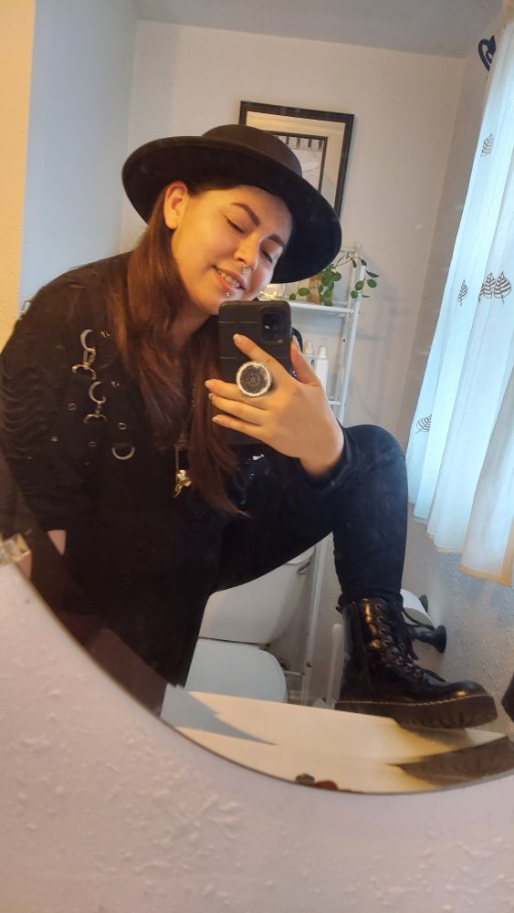 goth trans guy grinning at the camera. he's got long brown hair, 4 necklaces, a black wide brim hat and a spooky all black goth shirt with clips and buckles and stuff. he's got 1 boot up on the kitchen sink to show off their shiny black quality