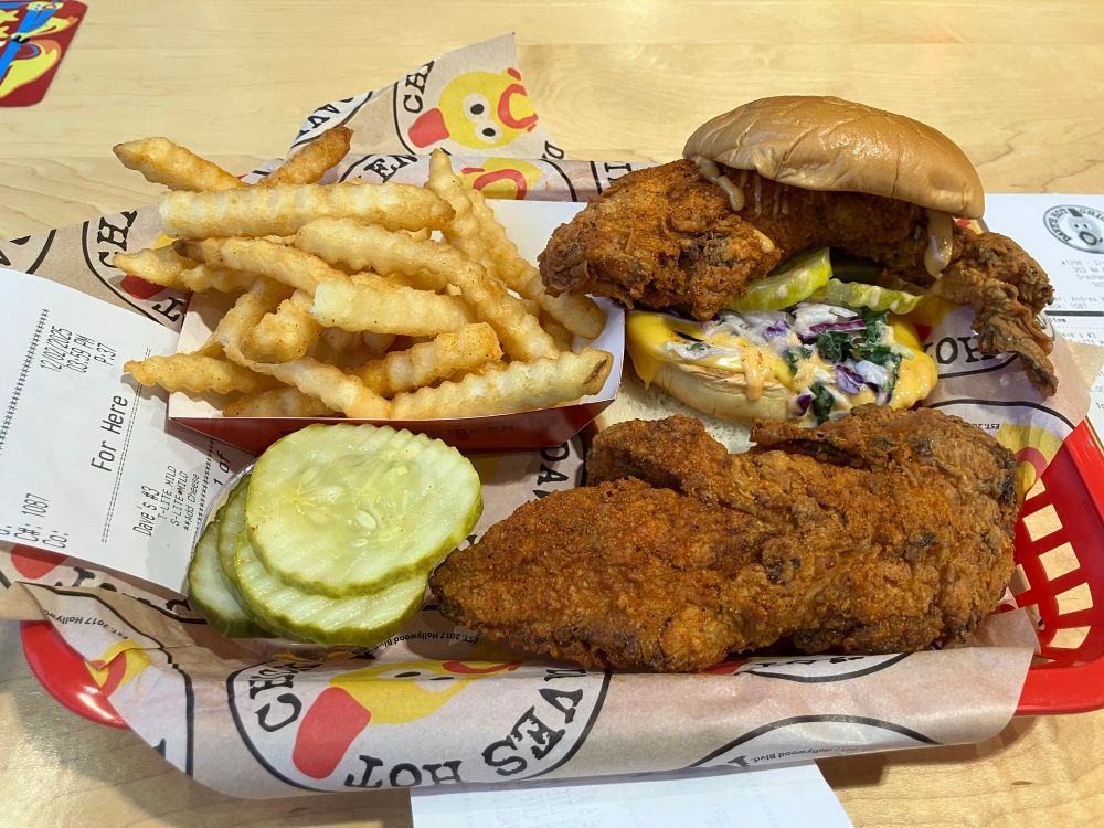 A picture of my tray of food from Dave’s Hot Kitchen. There is crinkle cut fries, some slices of pickles, a giant well cooked chicken tender, and a Dave’s slider, with Kale slaw and cheese.