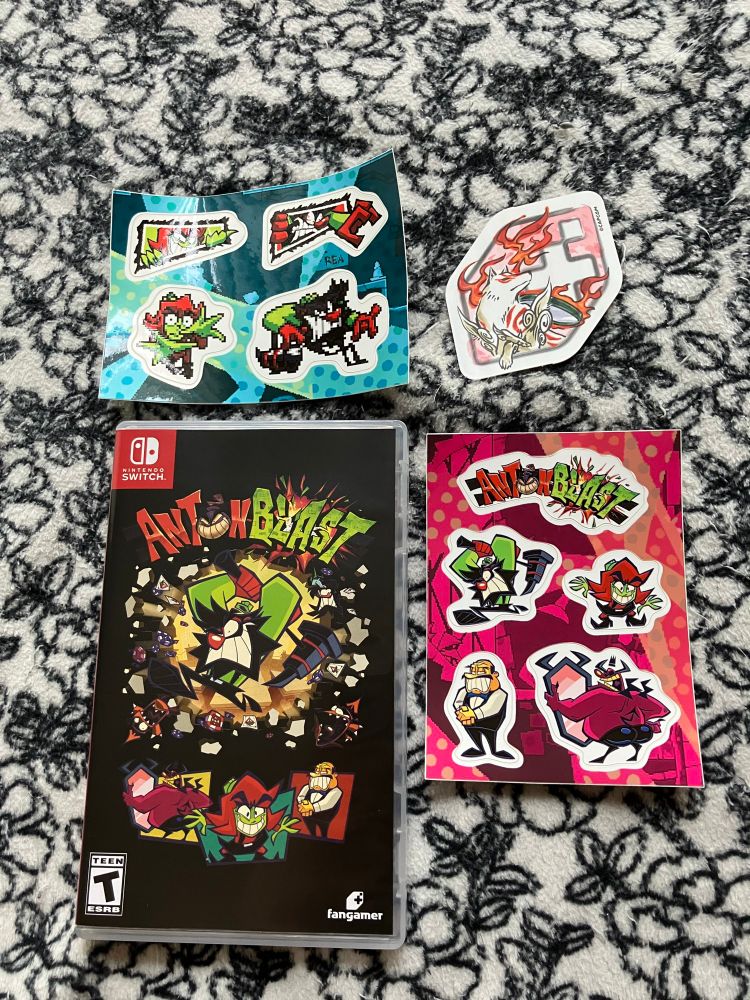 A physical copy of Antonblast for the Nintendo Switch, from Fangamer, on my bed. Included are two sticker sheets, and a Fangamer brand sticker with fanart of the game Okami.