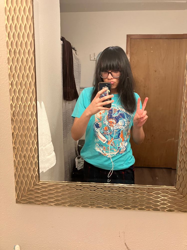 A selfie of myself. I’m standing in the bathroom, in front of the mirror. I’m wearing a teal t-shirt with a design of Lily from Street Fighter VI. I am holding up a peace sign, holding up my phone to take the photo.
