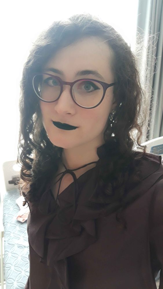 A selfie of Oceo, with long curly hair, reddish-purple glasses and matching eyeshadow, black lipstick and a dark purple frilly top.