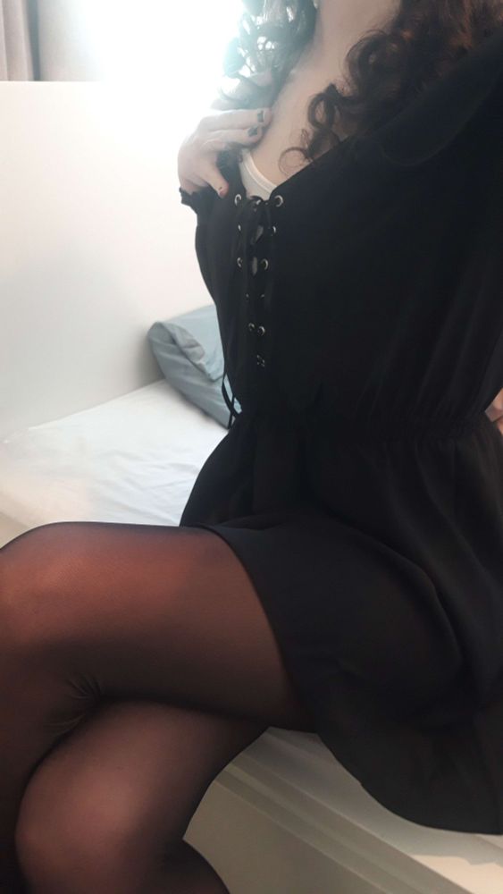 An Oceo selfie of her sitting down, showing her legs in sheer tights, her black dress and her curly hair.