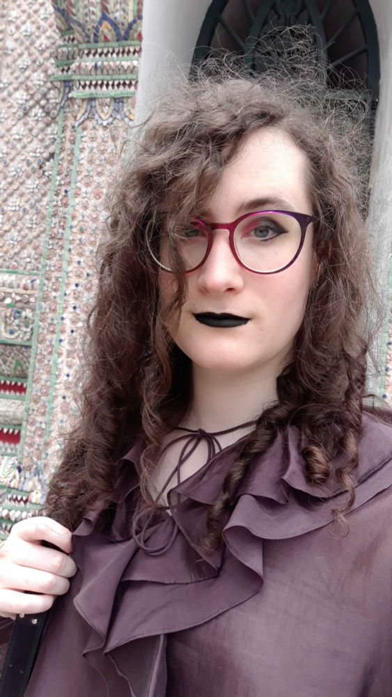 A selfie of Oceo, with long curly hair, reddish-purple glasses and matching eyeshadow, black lipstick and a dark purple frilly top. She's standing in front of an ornate mosaic building.