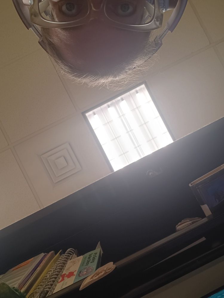 The tiniest bit of the top of my face is visible, wearing giant headphones. Mostly, the photo focuses on the ceiling at work and a bright light, with a big portion of the top shelf of my desk at work.