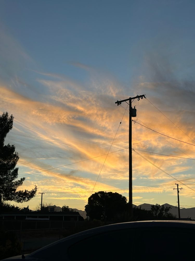 Sunset with orange and yellow tint in the clouds and blue sky. A silhouette of a telephone pole and trees can be seen in the foreground