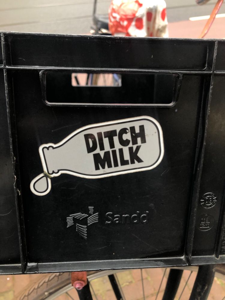 Picture of a sticker on the side of a black bike crate. The sticker is in the shape of a milk bottle and is white. In the middle, in black letters, it says ‘DITCH MILK’