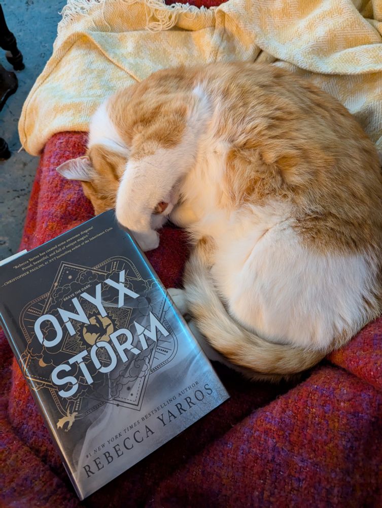 Orange and white cat sleeping on his side, leg covering his face, next to Jill's library copy of Onyx Storm.